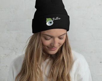 May include: Black beanie with a cartoon eye design wearing a green hat, and the name "Billie" embroidered in white. The beanie is made of a soft, knitted material.