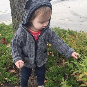 May include: A gray hooded sweater with black trim and bear ears. The sweater has a zipper closure and pockets. The child is wearing dark blue jeans and white shoes. The child is holding a rock and touching a plant.
