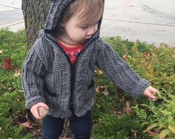 May include: A gray hooded sweater with black trim and bear ears. The sweater has a zipper closure and pockets. The child is wearing dark blue jeans and white shoes. The child is holding a rock and touching a plant.