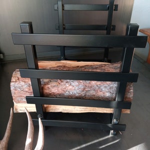 May include: Black metal log rack with a log of wood resting on the top. The rack has four horizontal bars and is standing on a grey surface.