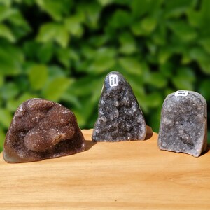 May include: Three amethyst geode clusters, two are gray and one is brown, all with a crystalline texture. The geode clusters are arranged on a wooden surface. The geode clusters are labeled "Made in Brazil".