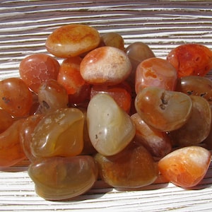 May include: A collection of carnelian tumbled stones in various shades of orange and brown. The stones are smooth and polished, with a natural, earthy look.