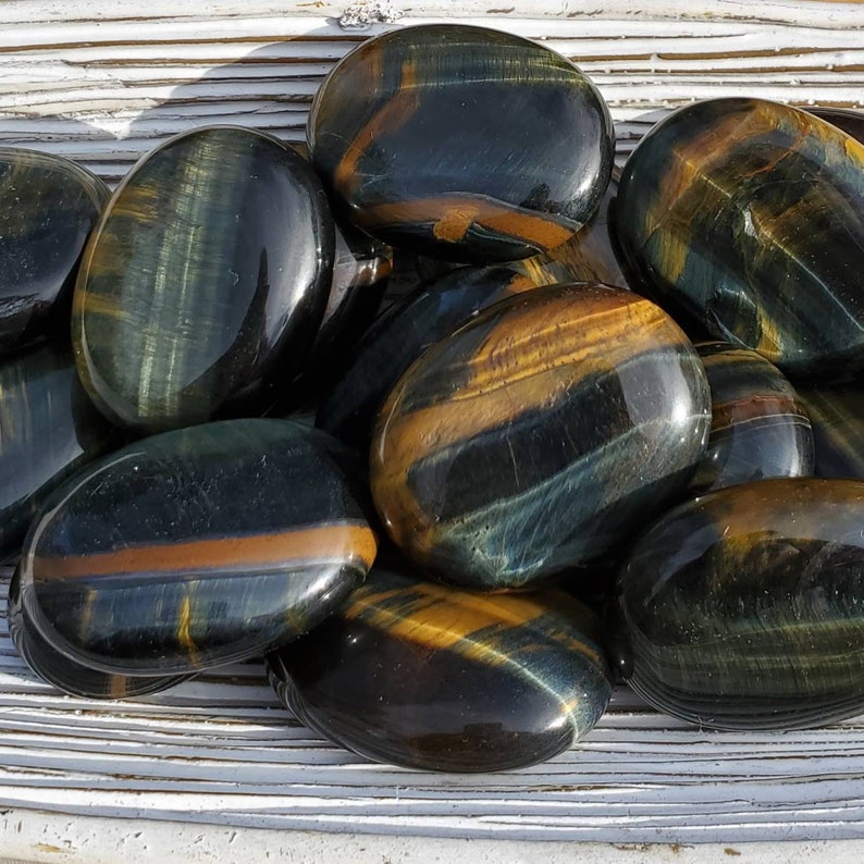 May include: A collection of smooth, oval-shaped blue tiger's eye stones. The stones have a dark blue base with golden brown stripes.