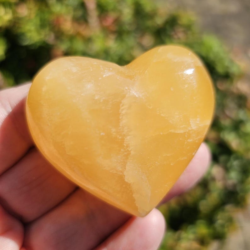 May include: A heart-shaped, polished, yellow calcite crystal. The crystal has a smooth, polished surface and a light yellow color.