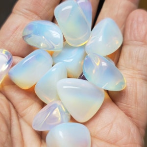 May include: A collection of polished opalite stones, each with a smooth, rounded shape. The stones display a milky white base color with iridescent flashes of blue, yellow, and pink. The stones are held in a hand.