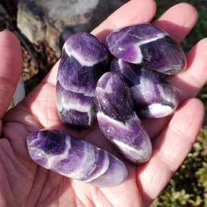 May include: A collection of polished amethyst stones, each displaying a rich purple hue with white banding. The stones are smooth and oval-shaped, held in a hand, showcasing their natural beauty and variations in color and pattern.