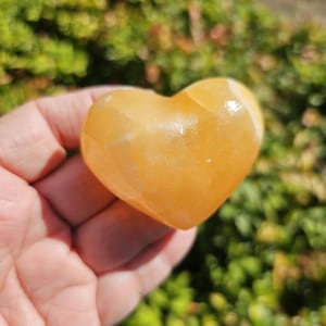 May include: A heart-shaped orange calcite crystal. The crystal is smooth and polished, with a light orange color. It is a natural stone and has a unique shape.