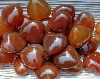 May include: A collection of heart-shaped carnelian crystals in various shades of orange and brown. The crystals are smooth and polished, with a glossy finish.