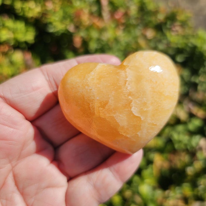 May include: A heart-shaped, polished, orange calcite crystal. The crystal has a smooth, polished surface and a warm, golden hue.