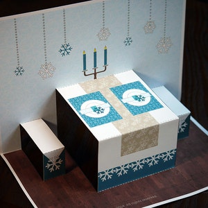 Puede incluir: Una tarjeta pop-up con un tema invernal. La tarjeta presenta una mesa con dos cubiertos, un candelabro con tres velas y un fondo azul con copos de nieve. La tarjeta est&aacute; abierta y la mesa est&aacute; levantada.