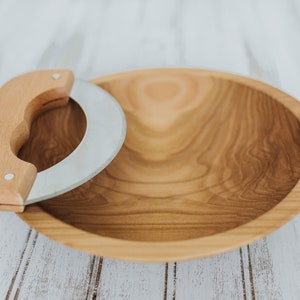 May include: A wooden bowl with a curved, stainless steel knife with a wooden handle. The bowl and knife are on a white wooden surface.