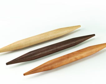 May include: Three wooden rolling pins, one light brown, one dark brown, and one light orange, are arranged in a row on a white surface.
