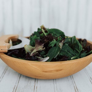 May include: A wooden salad bowl with a salad chopper inside. The bowl is filled with a variety of mixed greens.