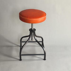 Könnte beinhalten: Ein brauner Lederschemel mit einem schwarzen Metallgestell. Der Schemel hat eine runde Sitzfläche und einen Höhenverstellmechanismus.