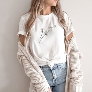 Pode incluir: Camiseta branca com um gráfico de duas sementes de dente-de-leão sendo levadas pelo vento. O texto "What will be will be" está escrito na camiseta.