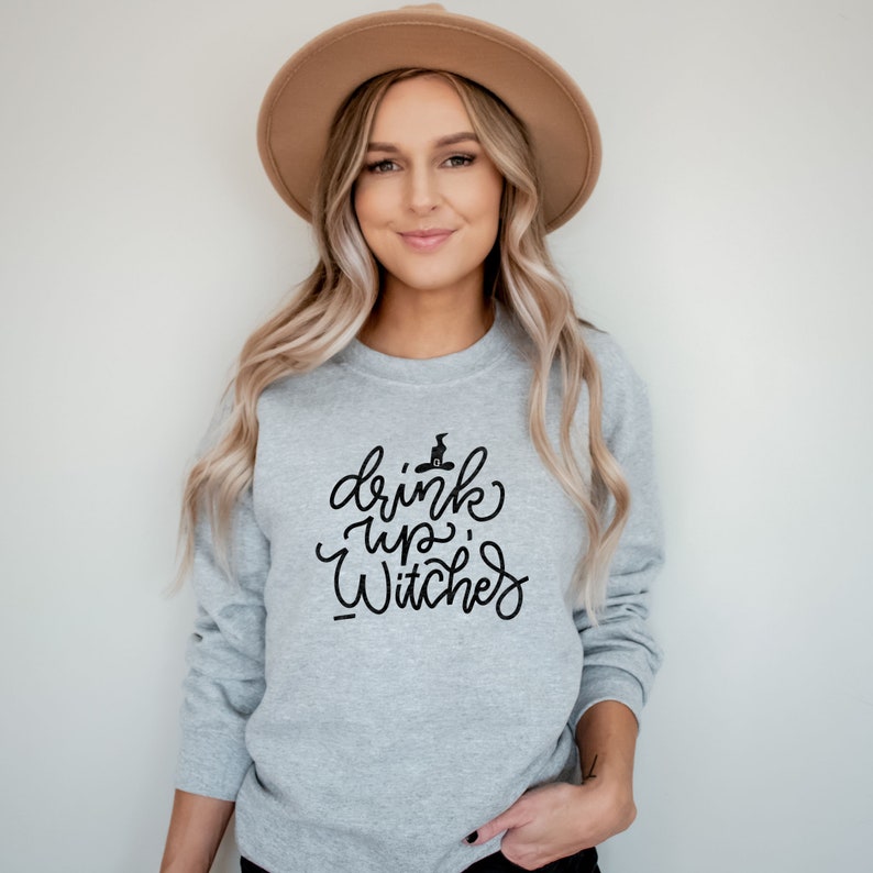 Puede incluir: Una sudadera gris con el texto "drink up, witches" en letra negra. El texto incluye un peque&ntilde;o sombrero de bruja negro con un gato en la parte superior.