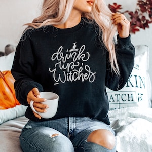 Puede incluir: Una sudadera negra con el texto "drink up, witches" en letras blancas. La sudadera tiene un gr&aacute;fico de sombrero de bruja sobre el texto.