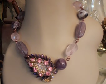 May include: A purple and pink beaded necklace with a large, sparkly, purple and pink brooch in the centre.