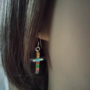 Peut inclure: Une paire de boucles d'oreilles en argent avec un pendentif en forme de croix colorée. La croix est en émail et présente un motif bleu, vert, orange et jaune.