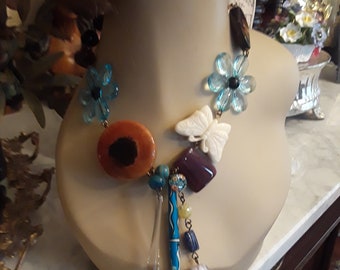 May include: A colorful statement necklace with blue flower accents, a butterfly charm, and a teardrop pendant. The necklace features a variety of beads, including turquoise, orange, and purple.