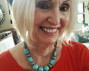 May include: A woman wearing a red shirt with a turquoise and green beaded necklace. The necklace features large, round turquoise beads.