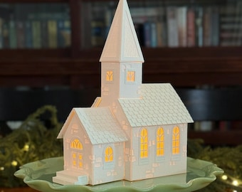 May include: A white church-shaped decorative light with a tall steeple and arched windows. The church sits on a light green cake stand. The windows glow with a warm light, suggesting an interior illumination. The overall design is detailed and elegant.