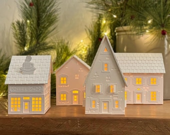 May include: Four white paper houses with lit windows, including a coffee shop with the text "Coffee Shop" on the front. The houses are on a wooden surface.