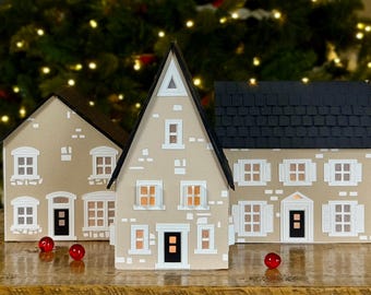 May include: Three miniature paper houses with white windows and doors, lit from within, on a wooden surface. The houses are decorated with a brick pattern and have black roofs. Two red ornaments are in the foreground.