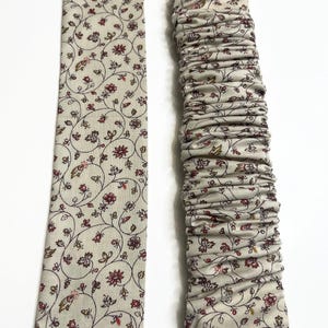 May include: A long fabric strip featuring a beige background with a repeating pattern of small red flowers and thin brown vines. One section of the fabric is laid flat and smooth, while the adjacent section is gathered into dense, horizontal ruffles.