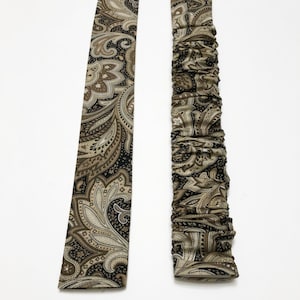 May include: Two fabric strips with a paisley pattern in shades of brown, tan, and black. One strip is flat, while the other is gathered, creating a ruched effect. The background is plain white.