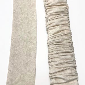 May include: Two beige fabric items with a paisley pattern. One is a long, flat strip, and the other is a gathered, ruched sleeve. Both items have a light beige background with a darker beige paisley design.
