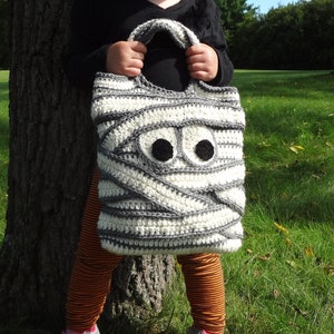 May include: A white and gray crocheted mummy-shaped trick-or-treat bag with black felt eyes.