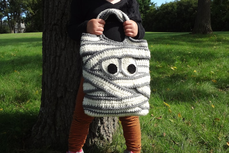 May include: A white and gray crocheted mummy-shaped trick-or-treat bag with black felt eyes.
