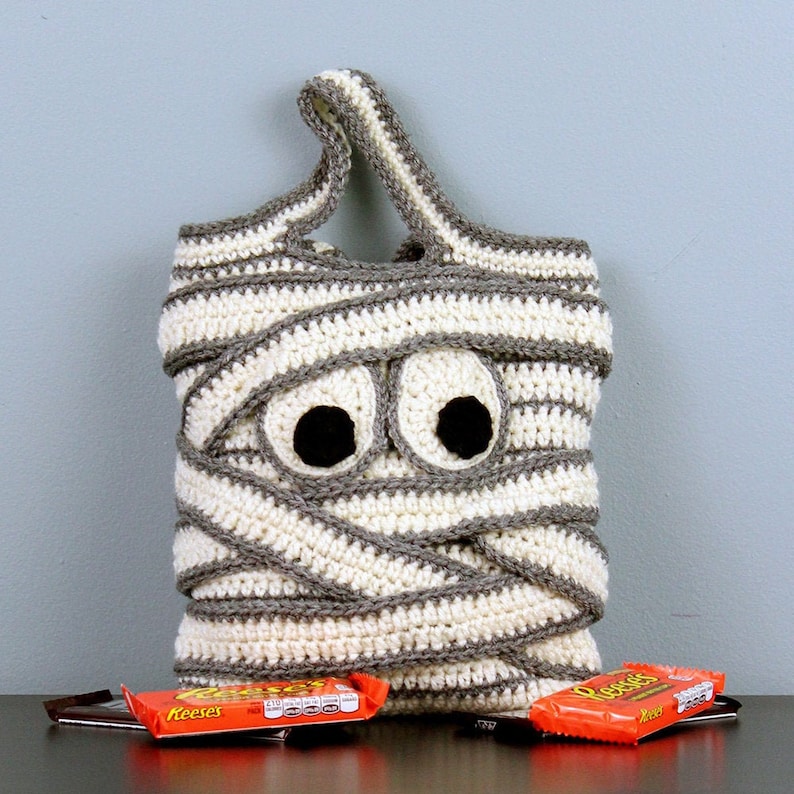 May include: A crocheted mummy-shaped trick-or-treat bag with googly eyes and a gray and white striped pattern. The bag is filled with candy bars.