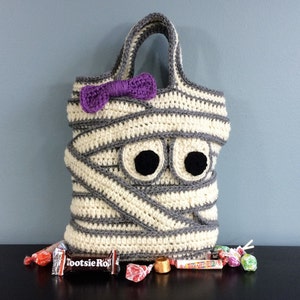 May include: A crocheted mummy costume bag with a purple bow, googly eyes, and the text "Tootsie Roll" on a candy bar in front of the bag.