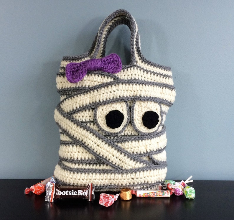May include: A crocheted mummy costume bag with a purple bow, googly eyes, and the text "Tootsie Roll" on a candy bar in front of the bag.