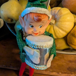 May include: A vintage Christmas elf figurine with a green glittery outfit, red pants, and black shoes. The elf is holding a silver drum with a white top and gold trim. The elf has red hair, blue eyes, and a smiling face. The background includes pears and pumpkins.