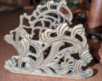 May include: Ornate brass napkin holder with a detailed, openwork design. The holder features scrolling patterns and floral motifs, showcasing a vintage aesthetic. The brass has a slightly aged patina, adding to its antique charm.