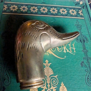 May include: A silver-colored duck head bottle stopper. The duck head is detailed with etched lines and a textured surface. The stopper has a brass-colored ring at the base. The background is a teal-colored book cover with gold floral designs.