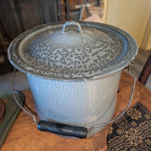 May include: A vintage, speckled gray enamelware bucket with a matching lid. The bucket has a wire handle with a black grip. The lid features a small handle. The bucket is sitting on a wooden surface.