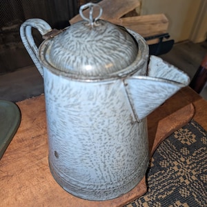 May include: A vintage enamelware coffee pot with a light blue and gray speckled pattern. The pot has a lid, handle, and spout. It is sitting on a wooden surface.