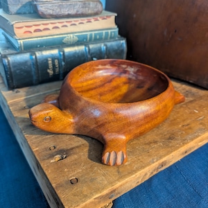 May include: A handcrafted wooden bowl in the shape of a seal. The bowl is carved from a light brown wood, with a smooth, polished finish. The seal has a detailed face and flippers. The bowl is sitting on a rustic wooden surface.