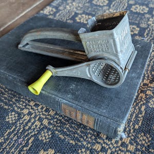 May include: Vintage metal rotary grater with a wooden handle and a yellow plastic handle. The grater has a rectangular hopper and a perforated grating surface. The text "U.S.A. PATENTS 2271175 2502867" is visible on the side. The grater is resting on a dark book.