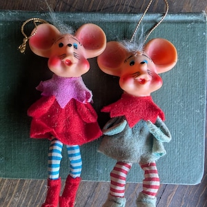 May include: Two hanging mouse ornaments with molded plastic heads, large ears, and painted facial features. One mouse wears a red felt outfit with blue and white striped legs, while the other wears a gray felt outfit with red and white striped legs.