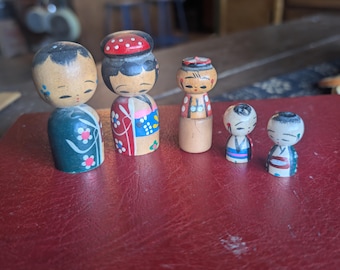 Könnte beinhalten: Eine Sammlung von sechs japanischen Kokeshi-Puppen. Die Puppen sind aus Holz gefertigt und mit verschiedenen Farben bemalt, darunter Rot, Blau und Grün. Die Puppen haben einfache, stilisierte Merkmale und sind auf einer roten Oberfläche angeordnet.