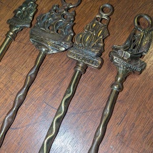 May include: Four antique brass fireplace tools with ship designs. The tools have long, twisted handles and decorative tops featuring sailing ships. One tool is labeled "Mayflower". The tools are arranged on a wooden surface.
