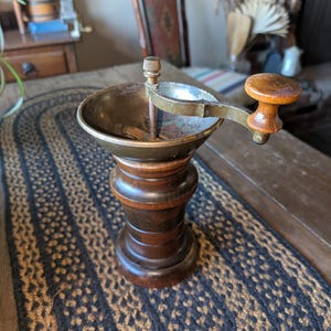 May include: A vintage wooden and brass coffee grinder with a crank handle. The grinder has a dark wood base and a brass top with a copper-colored interior. The crank handle is made of wood and has a brass knob.