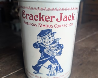 Puede incluir: Una lata cilíndrica de metal blanco con gráficos en rojo y azul. El texto dice Cracker Jack, America's Famous Confection y The More You Eat The More You Want. Una ilustración azul muestra a un marinero con cajas y un perro.