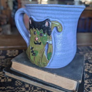 May include: A light blue ceramic mug featuring a raised, sculpted green cat figure with black hair, a stitched scar on its body, and a small pink nose. The mug has a curved handle and a smooth, glossy finish, showcasing a detailed, three-dimensional design.