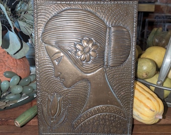 May include: A rectangular, bronze-colored metal relief sculpture featuring a woman's profile with a flower in her hair and a tulip. The artwork has a textured background and is framed with a detailed border. The piece is displayed on a wooden surface.